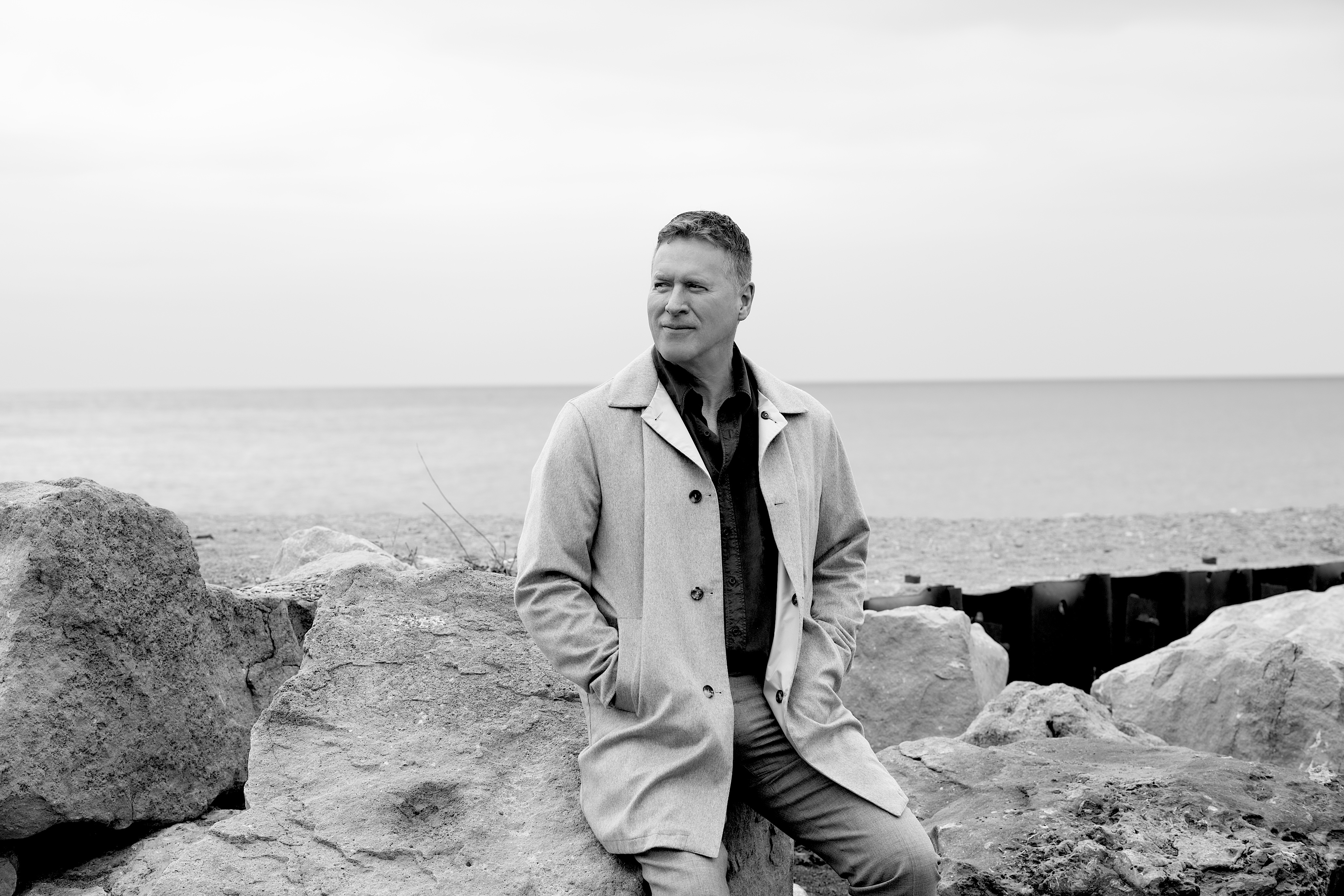 Black-and-white portrait of Ken seated on rocks beside the water.
