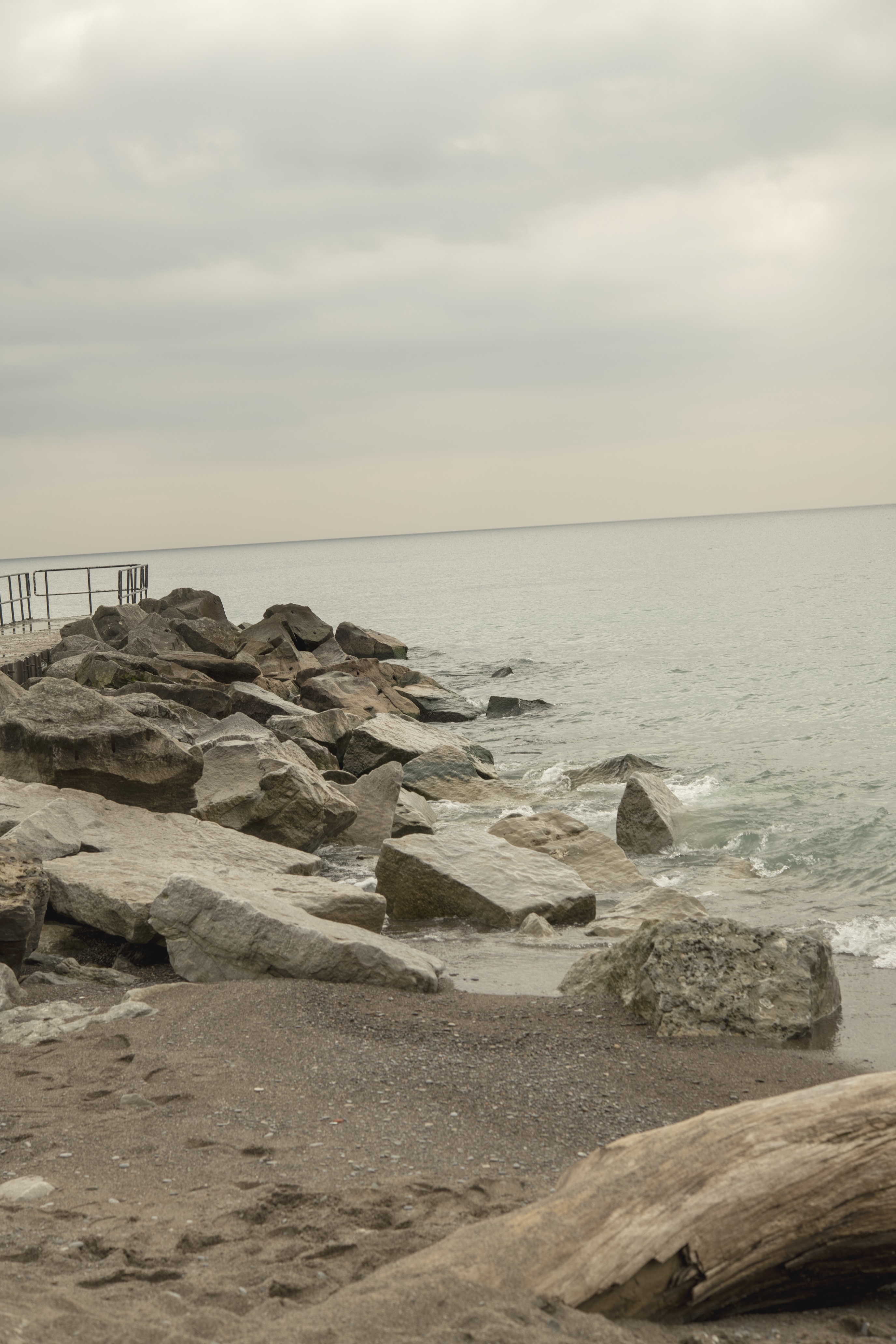 An open shoreline, with rocks, sand and water beneath a pale sky.
