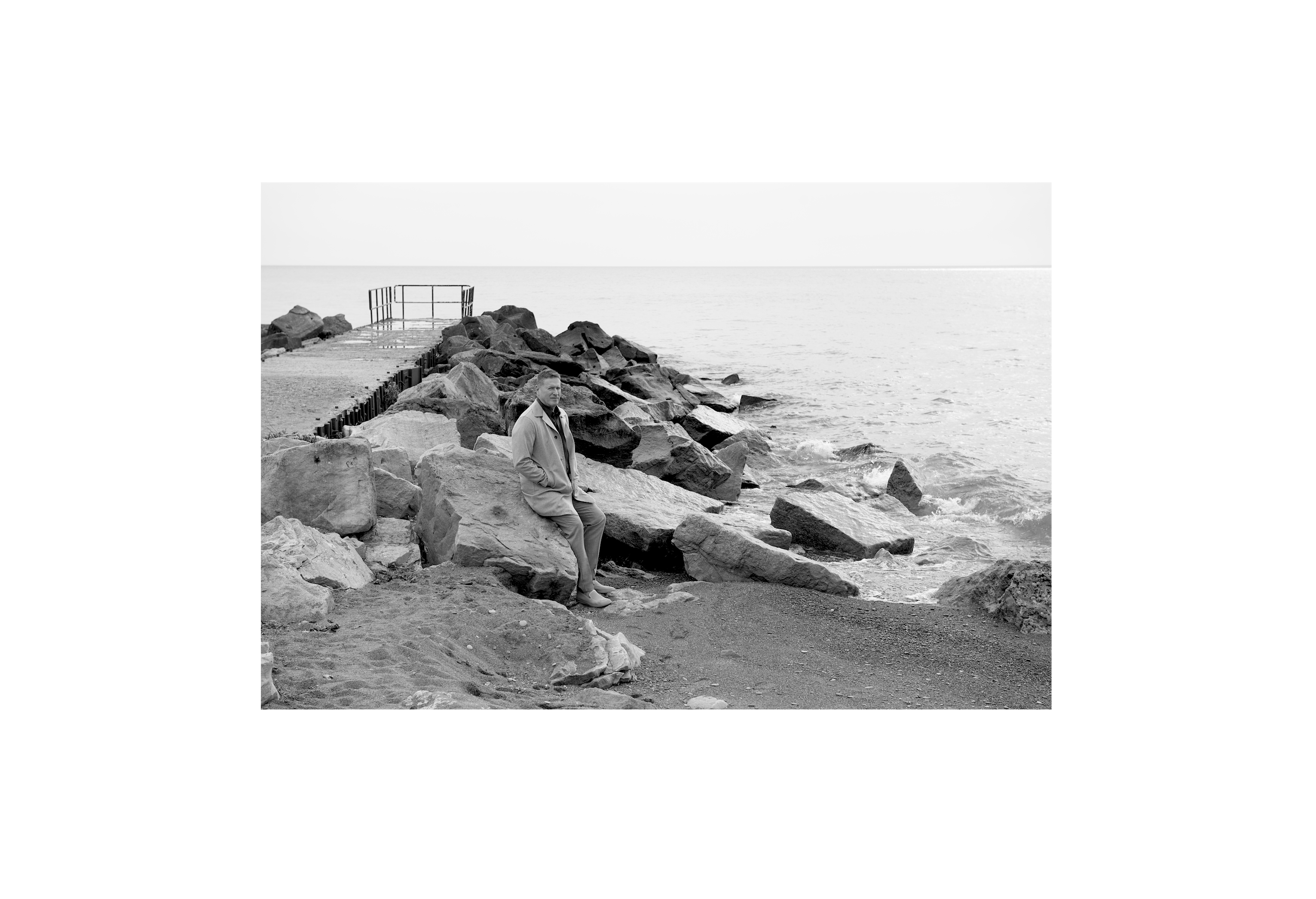 Ken Laird seated on rocks beside the water, photographed in black and white within a generous white border.